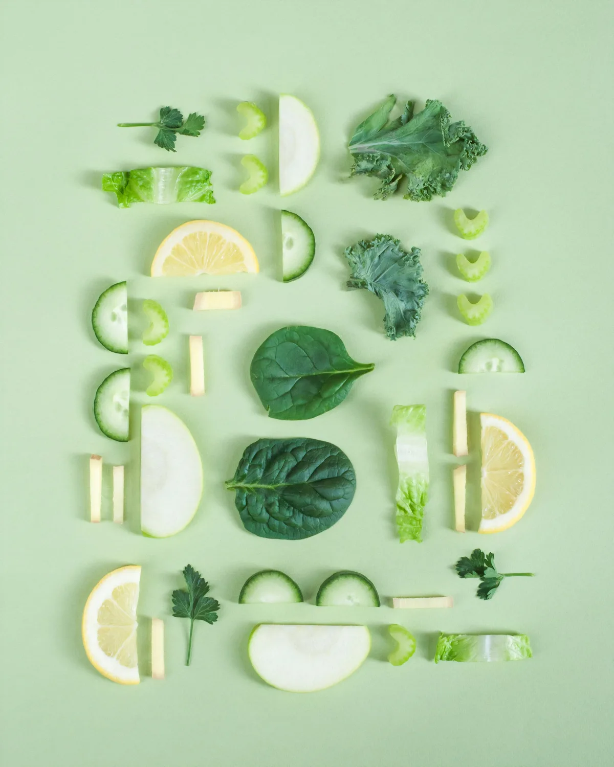 Composición ordenada de vegetales verdes frescos sobre fondo verde: espinaca, pepino, apio, kale y limón
